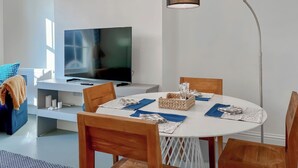 Ett modernt vardagsrum med en platt-TV, ett runt matbord med stolar, en golvlampa och en blå soffa med mönster.