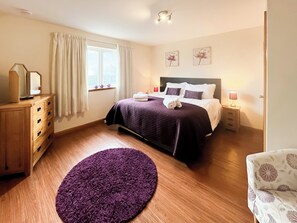Ein Schlafzimmer mit Bett, Kommode, Nachttisch, Lampe, Fenster und Vorhängen.