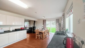 A modern kitchen with white cabinets, a dining area with wooden chairs, a black stove, and a large window with curtains. - 4 bedroom accommodation in Southerness (Southerness)