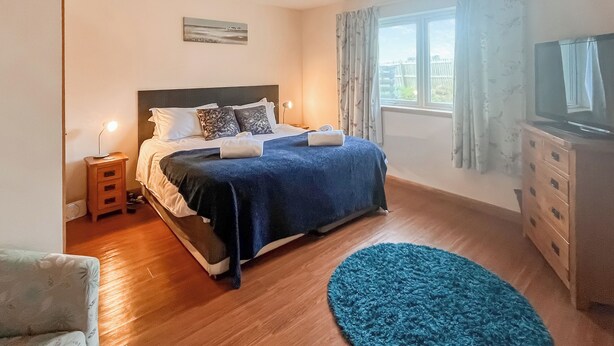 A bedroom with a bed, bedside table, lamp, TV, dresser, and a window with curtains. - 4 bedroom accommodation in Southerness (Southerness)