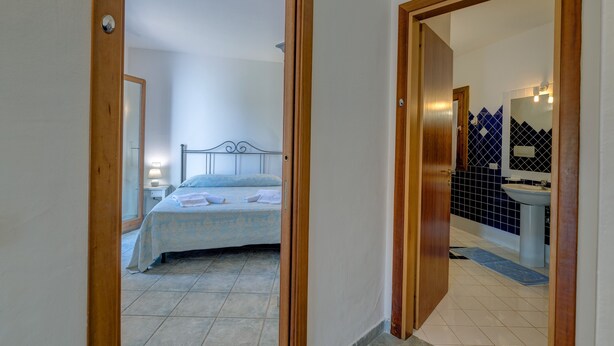 A bedroom with a bed, a nightstand, and a bathroom with a sink and mirror. - Casa Faro Green Village Sardegna (Palau)