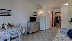 Ein kleiner, gut beleuchteter Raum mit Esstisch, gedeckten Weingläsern und einer Obstschale, einem Fernseher auf einer Holzkommode, einem weißen Kühlschrank und einer Couch mit gemustertem Bezug.