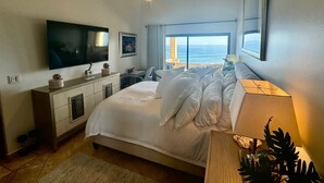 A bedroom with a bed, a TV, a desk, and a view of the ocean.