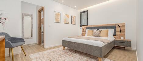 A modern bedroom with a grey bed, wooden headboard, and a bedside table with a lamp. A grey chair and a patterned rug are also visible.