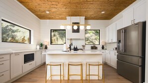 A modern kitchen with a wooden ceiling, white cabinetry, a central island with stools, and a stainless steel refrigerator. - Heavenly Falls Cabin in Broken Bow | Waterfall Retreat (Broken Bow)