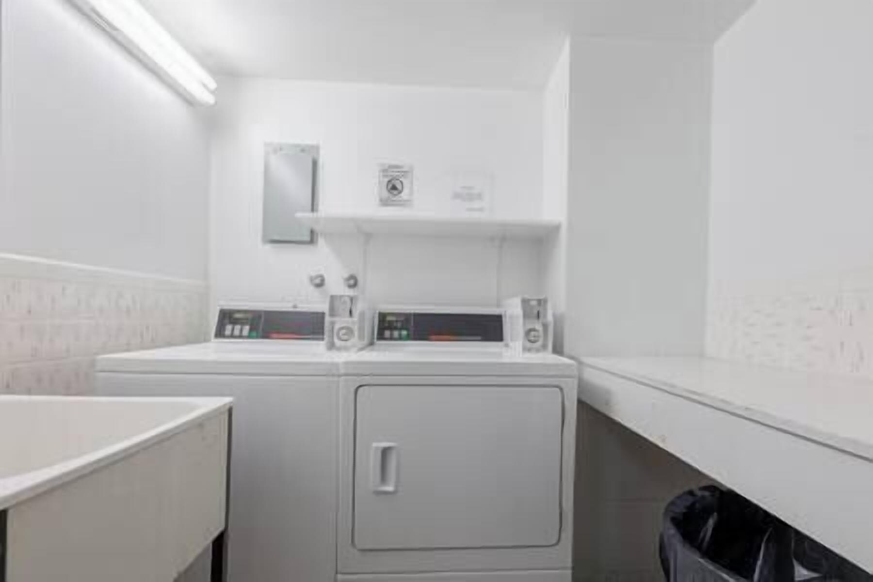 Een wasserette met een voorlader wasmachine en droger, een prullenbak en een plankje met een klein voorwerp erop.