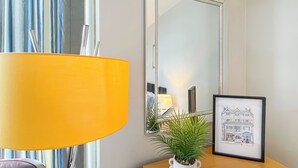 Een kamer met een houten bureau, een gele lamp, een ingelijste afbeelding en een potplant.