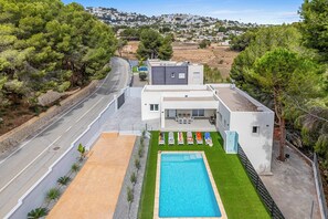 A modern house with a swimming pool, a road, and a view of a town in the background. - Villa Aiko (Teulada)