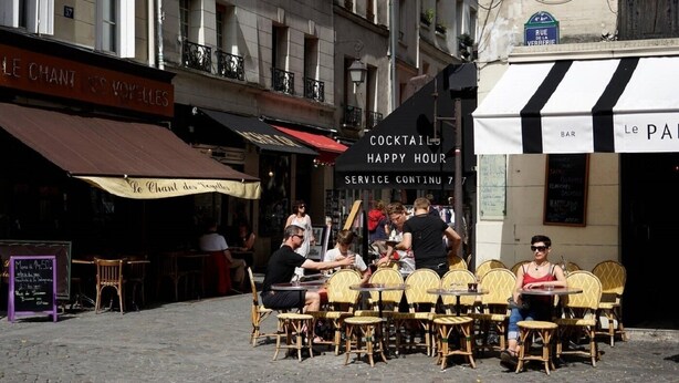 An outdoor café with wicker chairs and tables, a sign for 'Le Chant des Volettes', and a 'COCKTAIL HAPPY HOUR' banner. - A TRUE PARISIAN DUPLEX IN THE HEART OF PARIS - MARAIS - RUE DE LA VERRERIE (Paris)