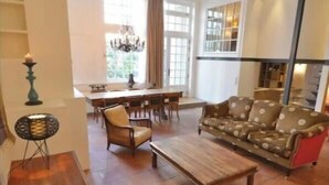 A spacious living room with a wooden coffee table, a patterned sofa, and a red rug. There is a dining area with a chandelier and a window with curtains. - A TRUE PARISIAN DUPLEX IN THE HEART OF PARIS - MARAIS - RUE DE LA VERRERIE (Paris)