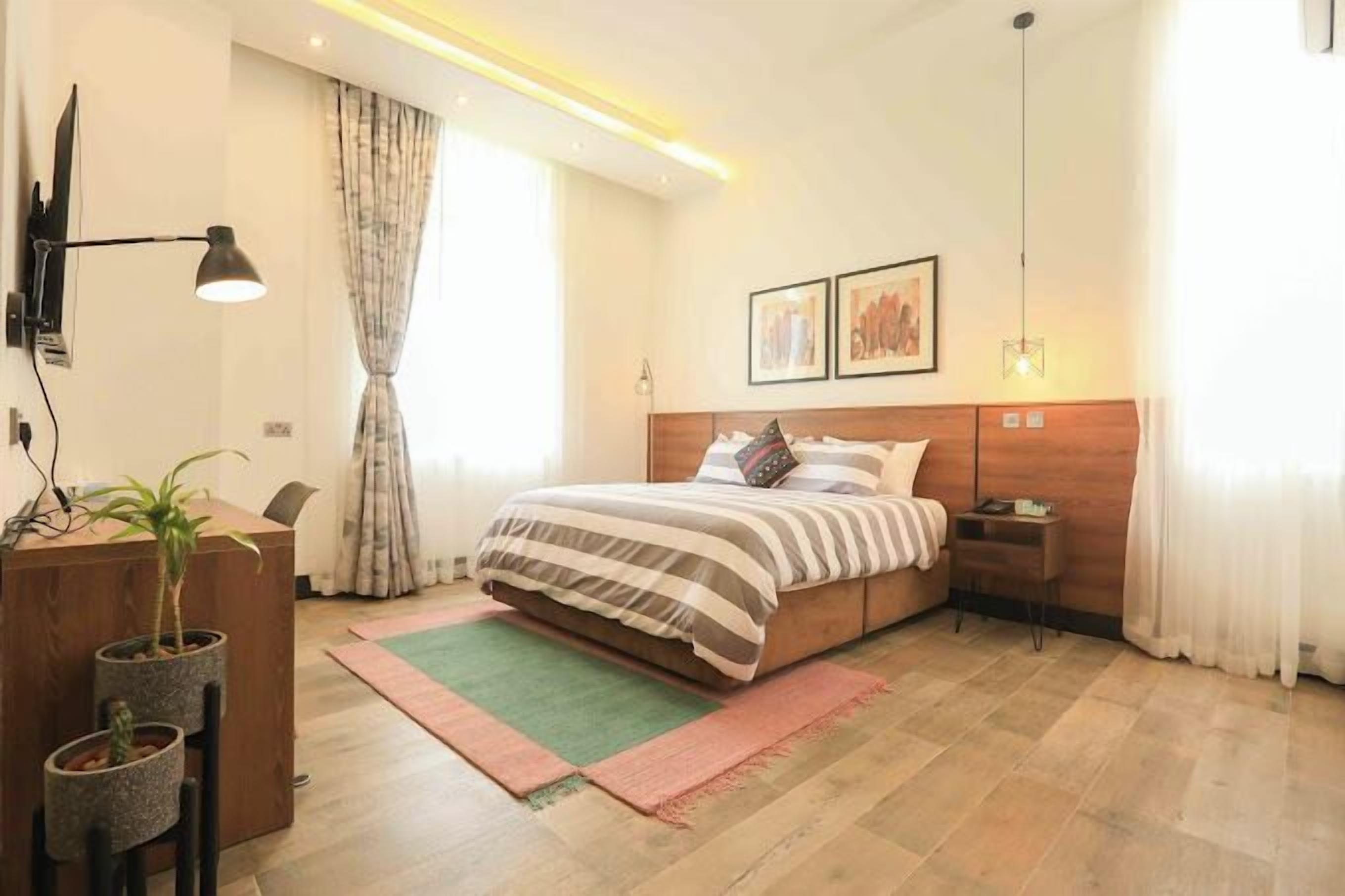 A bedroom with a bed, bedside tables, a desk with a lamp, a potted plant, and a window with curtains.