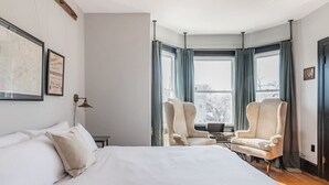 Une chambre d’hôtel avec un grand lit, deux fauteuils, une petite table et une fenêtre avec des rideaux.