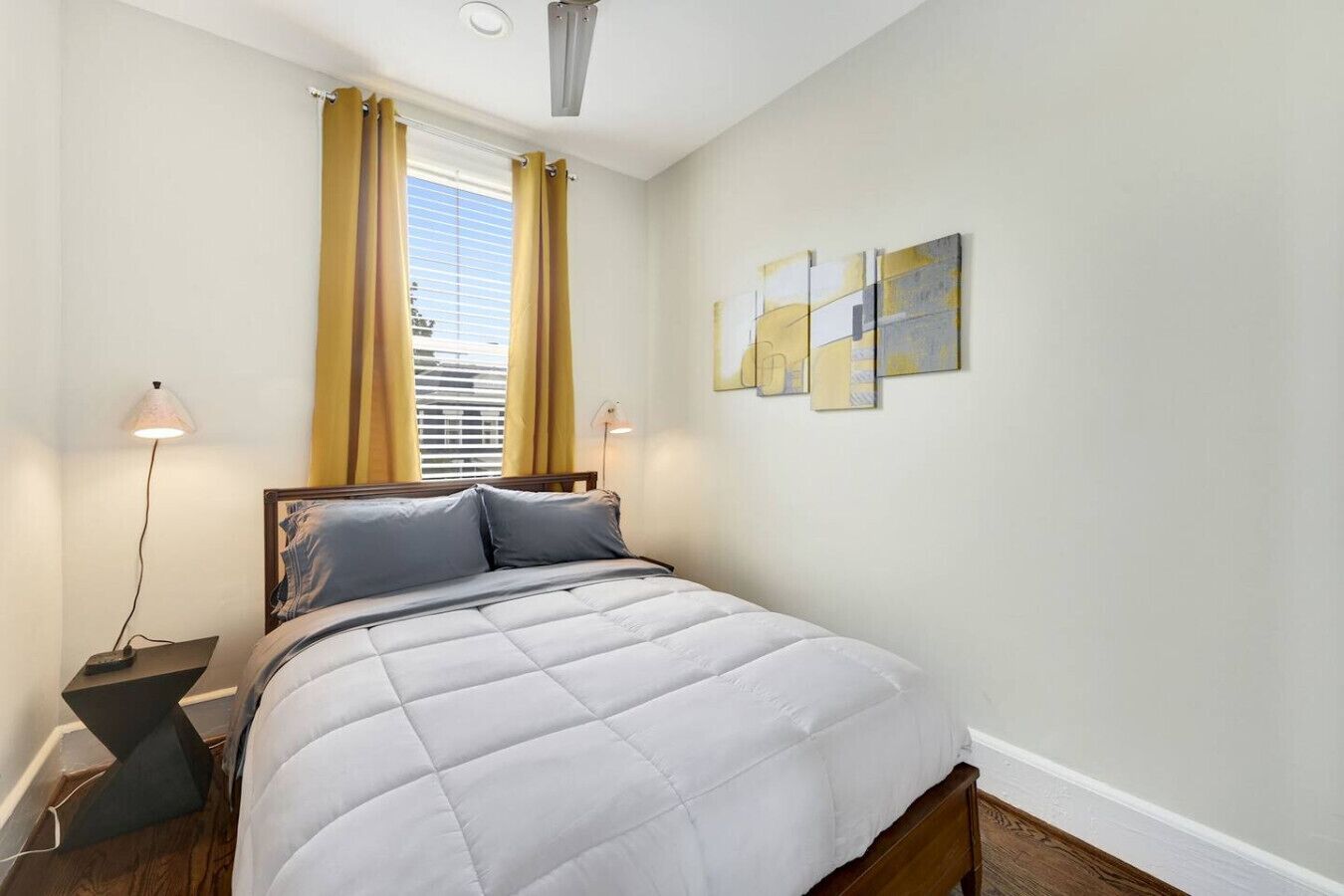 A bedroom with a bed, a window with curtains, a bedside table with a lamp, and wall art.