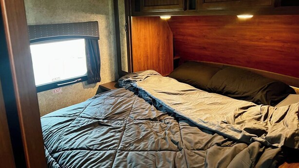 A compact RV interior with a bed, wooden cabinets, and a window with an exit sign. - Ample Lakeside Travel Trailer (Bridgeport)