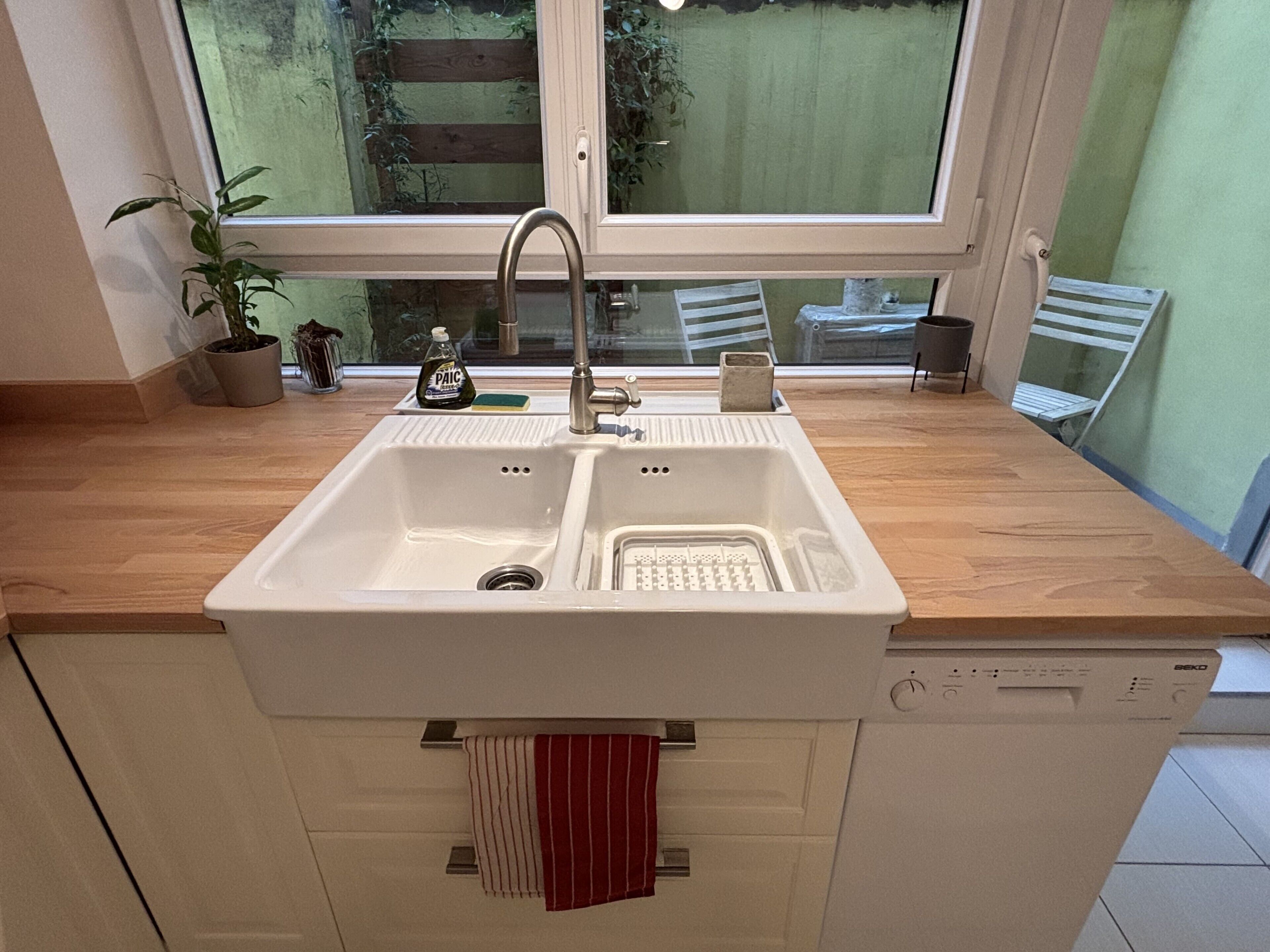 A kitchen with a double sink, wooden countertop, and a window with a view of greenery outside.