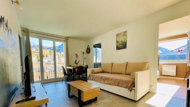 A modern living room with a sofa, coffee table, and TV stand. There is a dining area with chairs and a table. The room has large windows with a view of mountains. - Charming T3 with balcony in the heart of Briançon (Briançon)