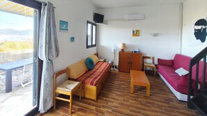 Un espacio habitable compacto con sofá, cama, mesa de madera y silla. Cuenta con ventana con cortinas, aire acondicionado de pared y vistas al exterior.
