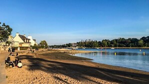 A lakeside area with a sandy beach, a few people, and buildings in the background. - Charming T3 with balcony near beach (Vannes)