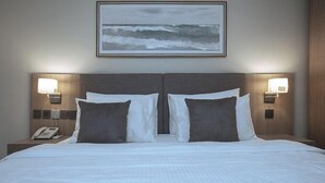 A hotel room with a large bed, two bedside lamps, a phone on the nightstand, and a painting of the ocean on the wall.