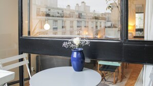 A room with a round table, a blue vase with flowers, and a view of buildings through a window.