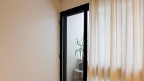 A room with a bed, a window with sheer curtains, and a view of a plant outside.