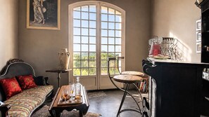 A room with a bed, a table with drinks, a chair, a large window, and a bookshelf.