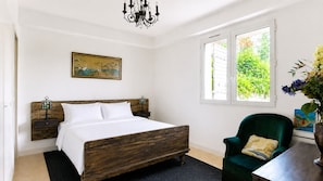 A bedroom with a wooden bed, a green armchair, a painting on the wall, and a window with a view of greenery.