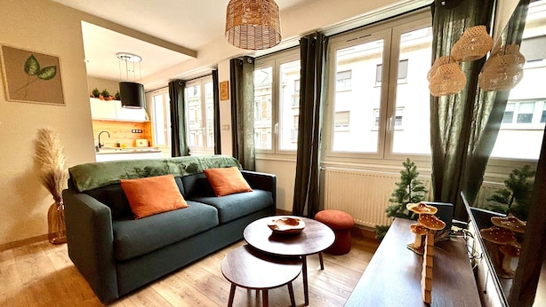 Un salon moderne avec un canapé, une table basse ronde et une table à manger. De grandes fenêtres, ornées de rideaux, laissent entrer la lumière, et un luminaire suspendu illumine l’espace. On peut admirer la vue sur la ville.