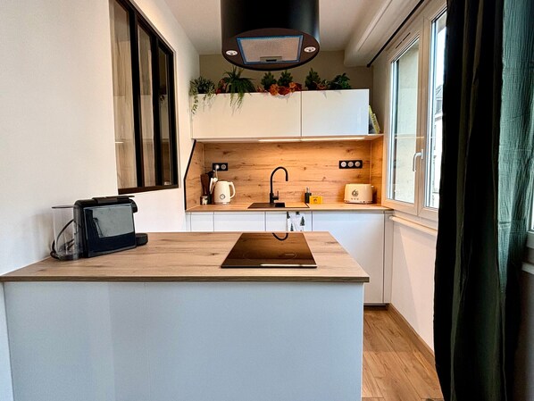 A modern kitchen with a central island, wooden cabinets, a built-in stove, and a window with curtains. - Nature Cocoon - Strasbourg Les Halles (Strasbourg)