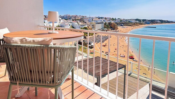 Un balcon avec une table et une chaise, donnant sur une plage parsemée de parasols et où se trouvent des gens.