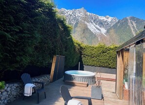 A hot tub on a wooden deck with chairs, surrounded by greenery and a mountain view.