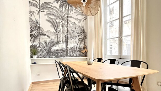 Een eetruimte met een houten tafel en zwarte stoelen, een groot raam met uitzicht op gebouwen, en een muurschildering van tropisch groen.