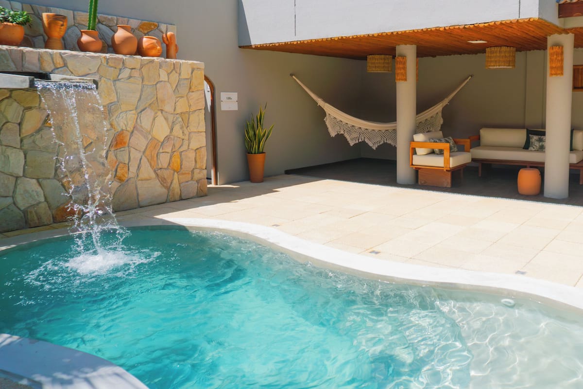 A swimming pool with a waterfall feature, surrounded by a stone wall and potted plants. There is a hammock and outdoor seating area with cushions.