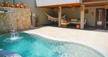 A swimming pool with a waterfall feature, surrounded by a stone wall and potted plants. There is a hammock and outdoor seating area with cushions.