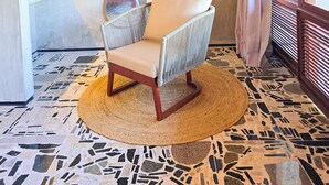 Una silla de mimbre con cojín, un tapete redondo tejido, techo de madera, lámpara de pared y una ventana con cortinas.
