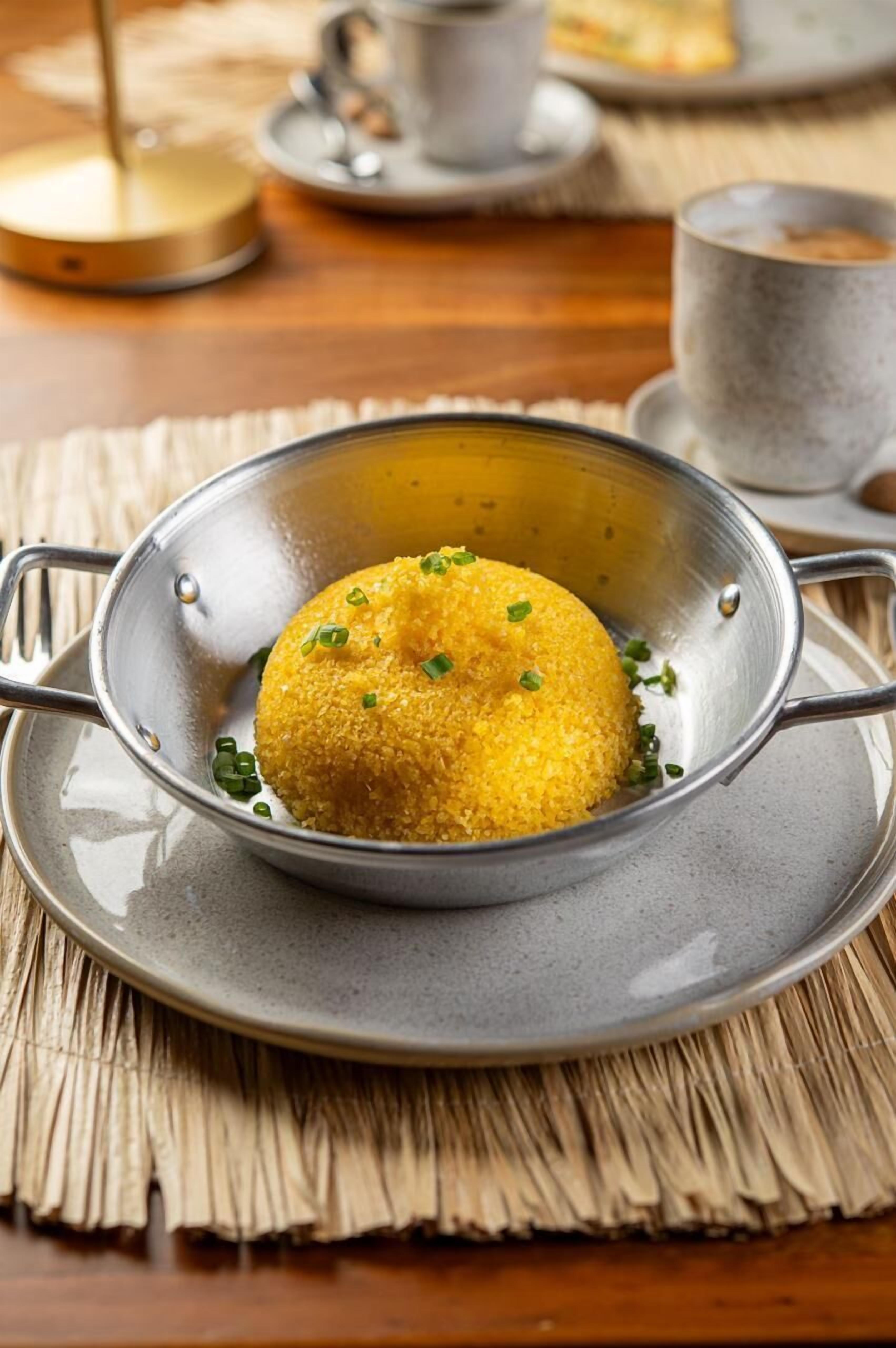 A metal colander containing a yellow food item garnished with green herbs, placed on a gray plate with a woven placemat underneath.