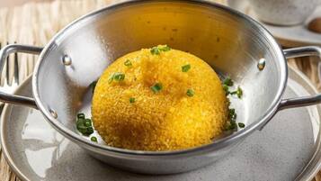 A metal colander containing a yellow food item garnished with green herbs, placed on a gray plate with a woven placemat underneath.