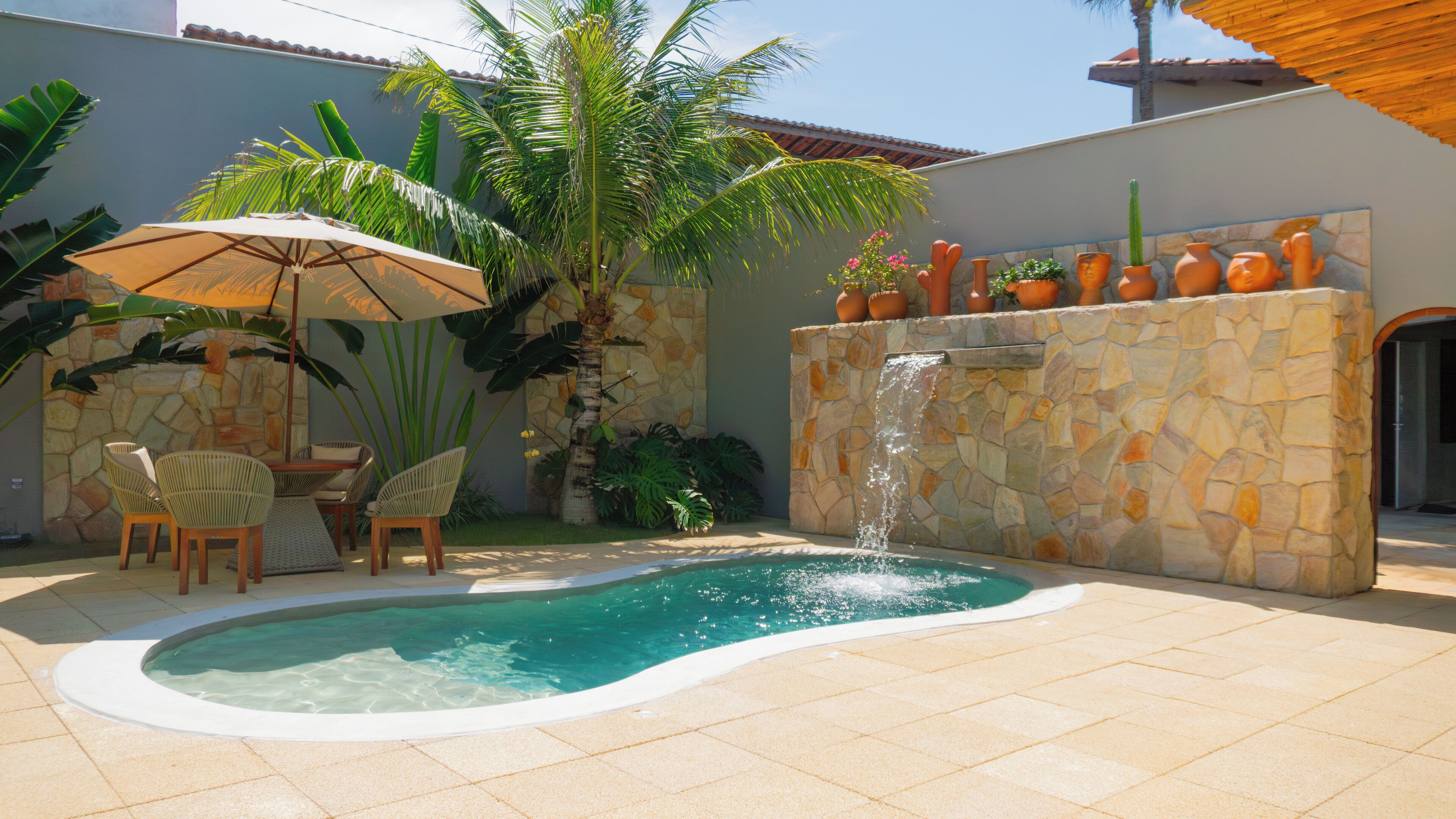 Una piscina con pared de piedra y una cascada, rodeada de sillas y plantas en macetas.