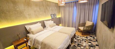 Quarto de hotel moderno com uma cama grande, duas mesas de cabeceira, uma cadeira, uma televisão e um chão estampado.
