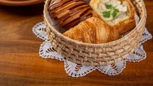 Una cesta de mimbre con croissants y un pastel, un plato con comida y un vaso de bebida roja sobre una mesa de madera.