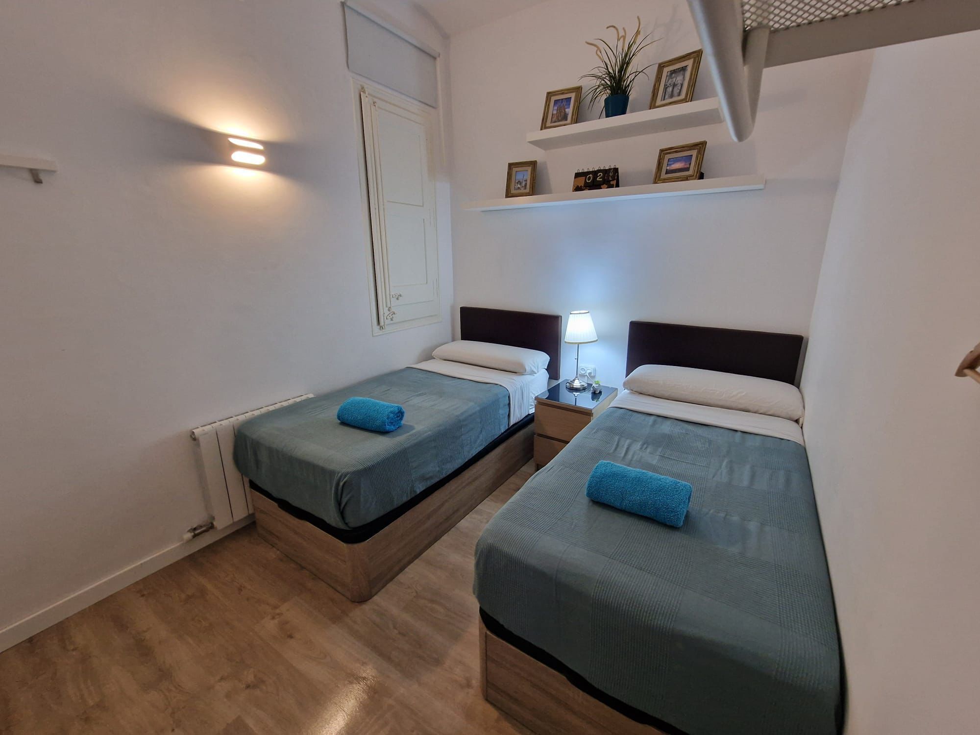 Hotelkamer met twee enkele bedden, een nachtkastje met lamp, een plank met ingelijste foto’s en een agenda, en een raam met jaloezieën.