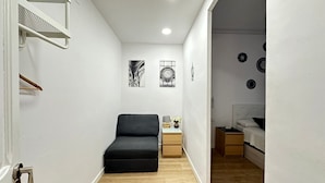 A narrow hallway leading to a bedroom with a bed, a bedside table, and a chair. There are two wall-mounted shelves and a door with a handle. - Rooms Flor (Barcelona)