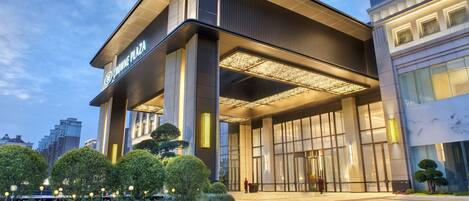 A modern hotel entrance with a large glass facade, a well-lit entrance area, and a landscaped garden with neatly trimmed bushes.