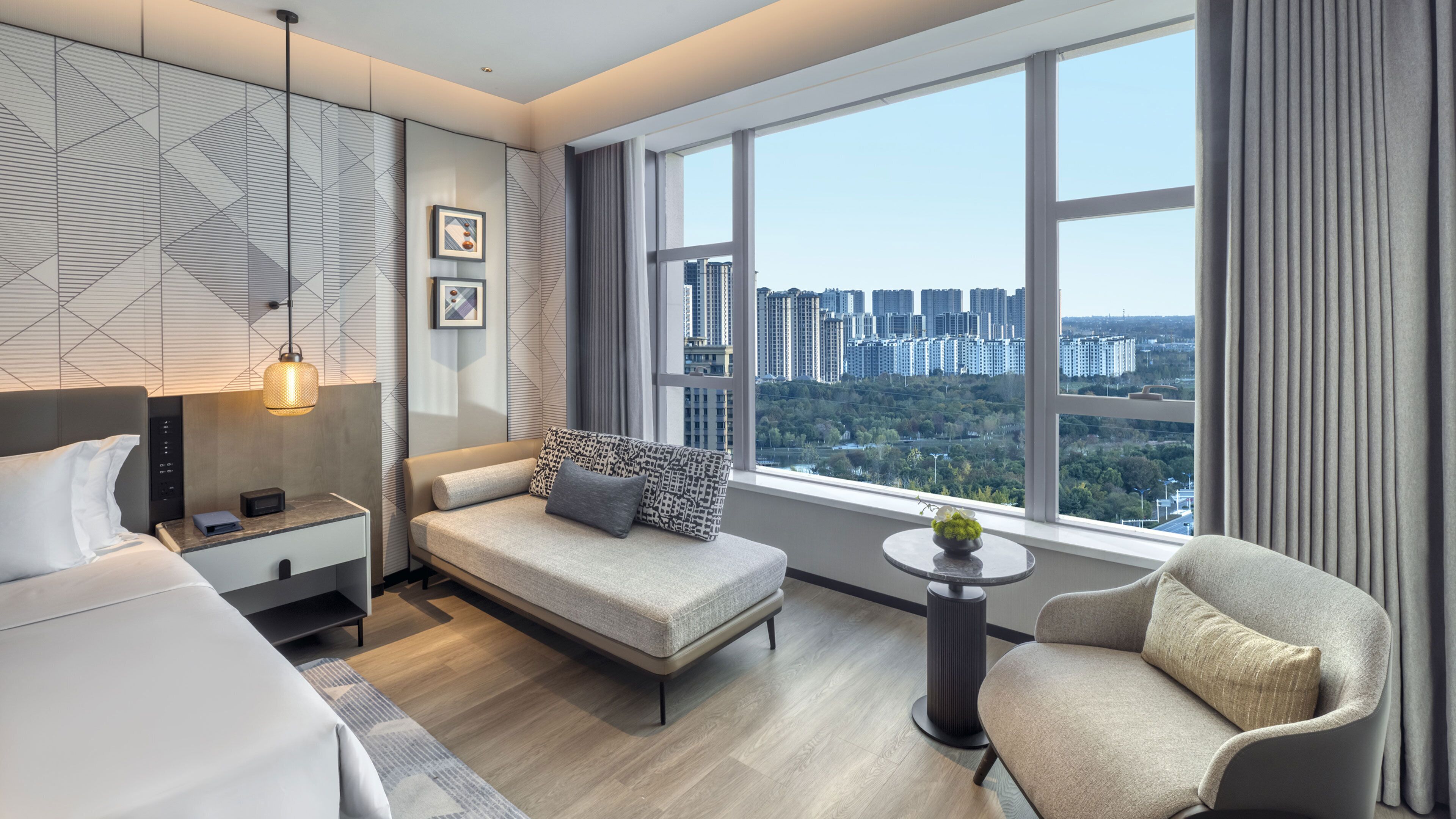 A modern hotel room with a bed, a sofa, a chair, a small table with a vase, and a large window offering a city view.