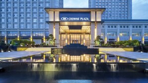 Het exterieur van het Crowne Plaza hotel met een reflecterend waterpartij ervoor, omringd door verzorgde beplanting en moderne architectuur.