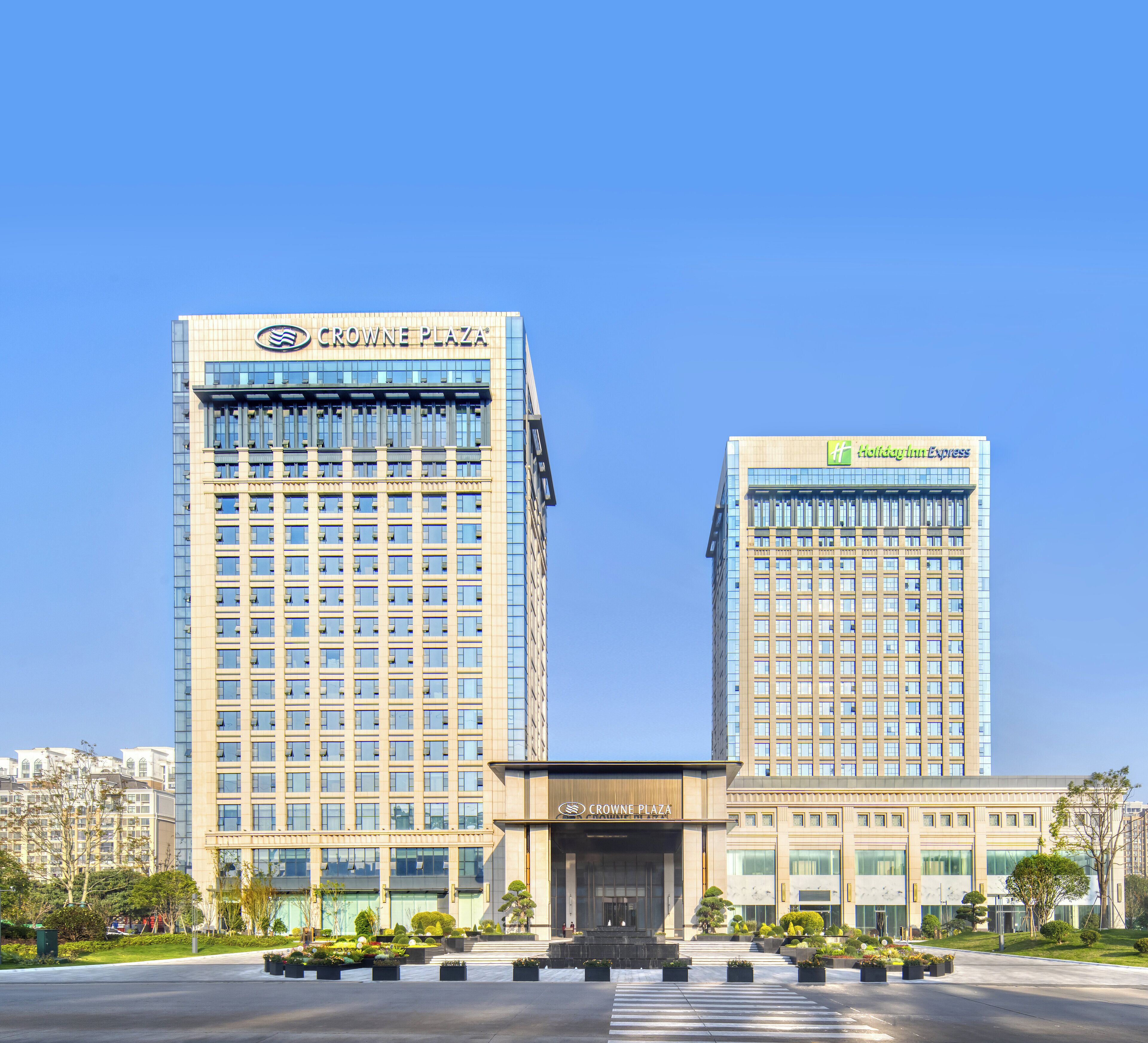 Two high-rise buildings with "Crowne Plaza" and "Holiday Inn Express" signage, surrounded by landscaped gardens and a clear sky.