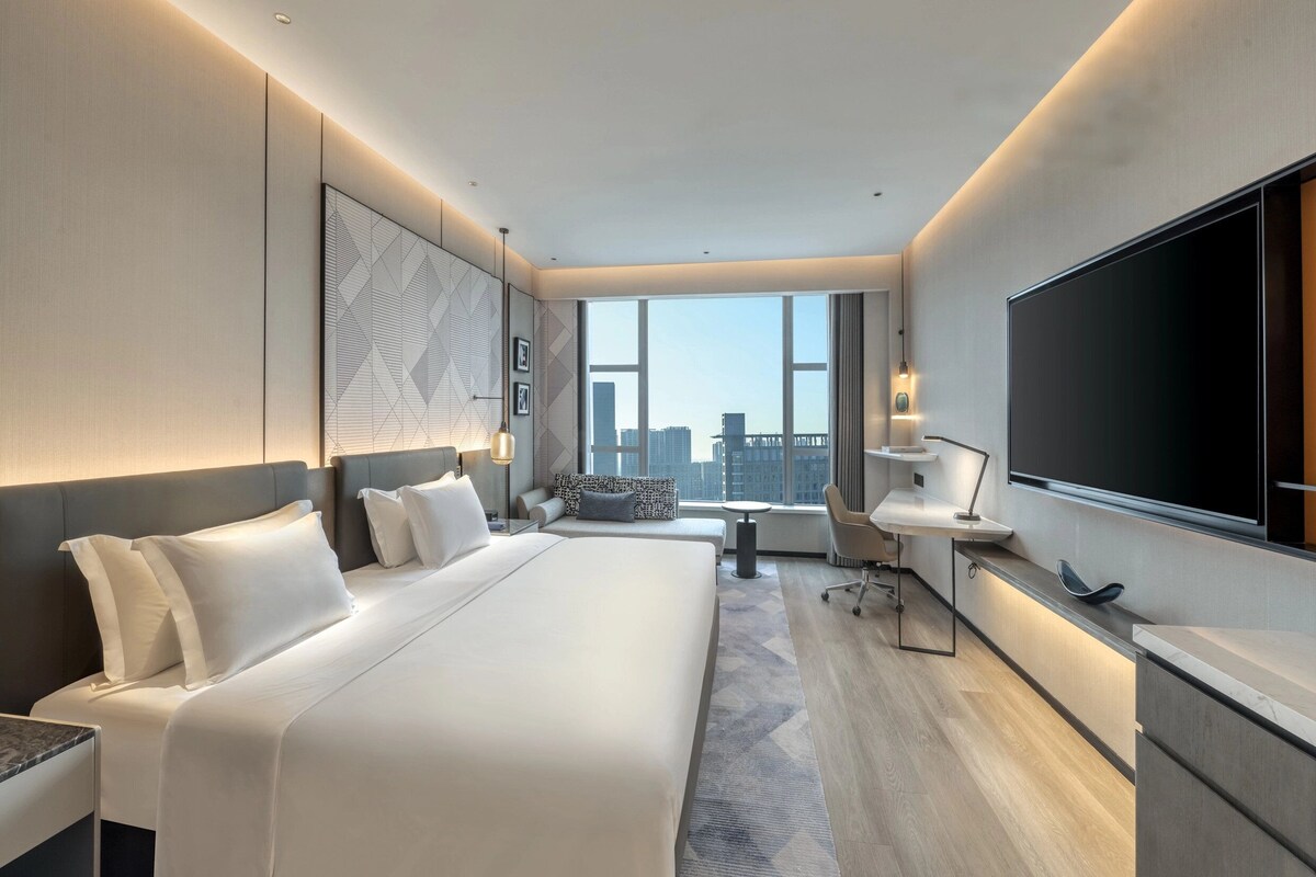 A modern hotel room with a large bed, a desk, a chair, a TV, and a city view.