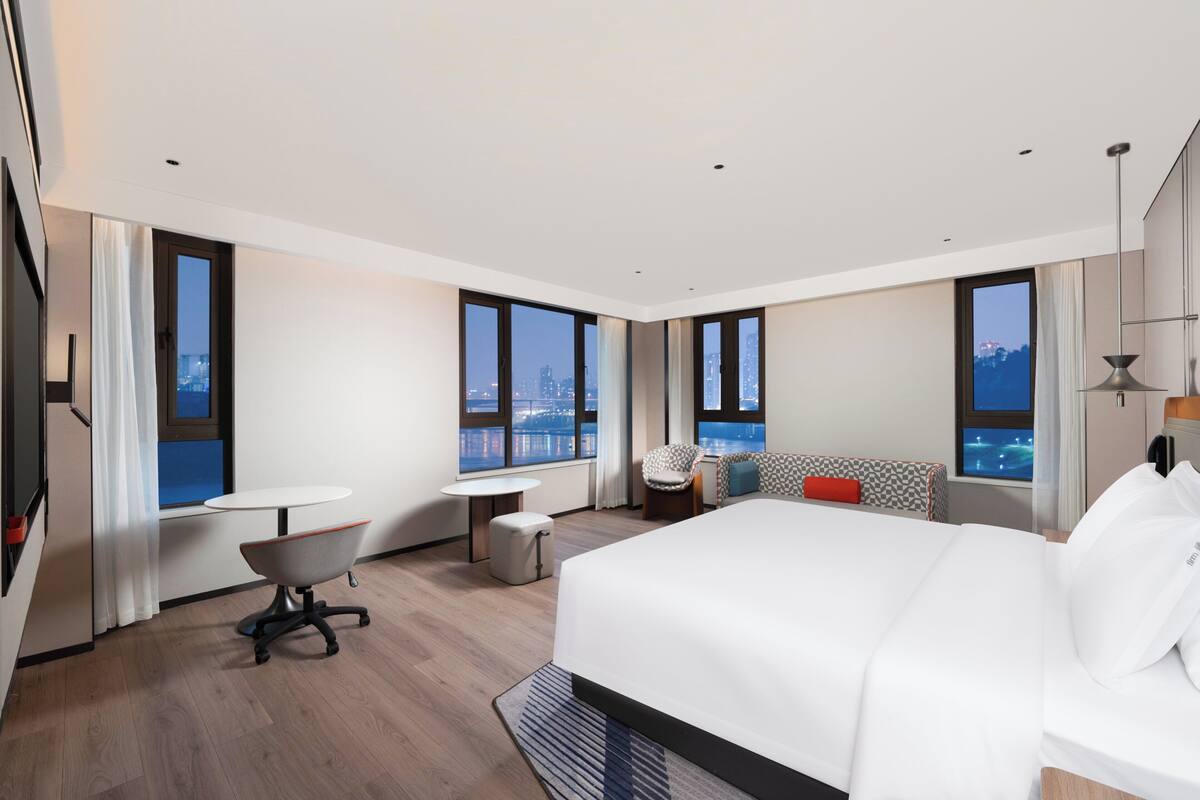 Ein modernes Hotelzimmer mit einem großen Bett, einem Schreibtisch, einem Sessel und einer Couch. Große Fenster bieten Blick auf die Stadt.