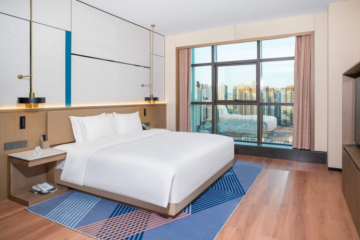 Habitación de hotel moderna con una cama grande, una mesita de noche, un escritorio y vistas a la ciudad a través de amplias ventanas.