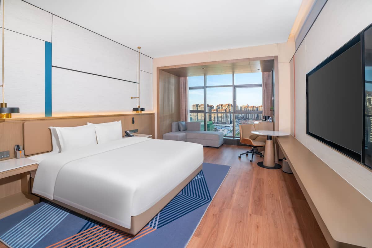 Habitación de hotel moderna con una cama grande, un escritorio, una silla, un televisor y vistas a la ciudad.